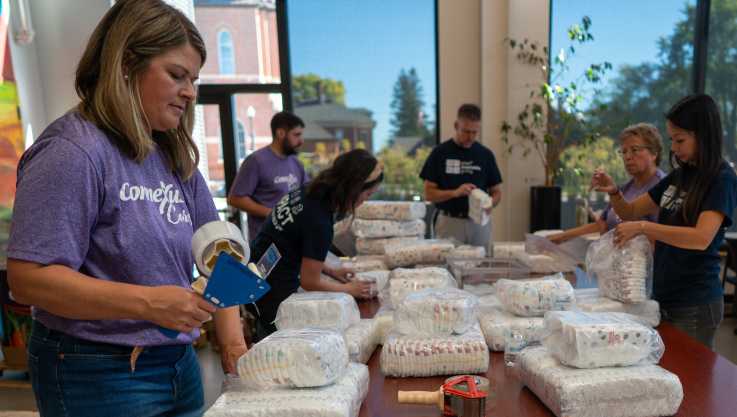 Six volunteers sort and package diapers at a large table. 