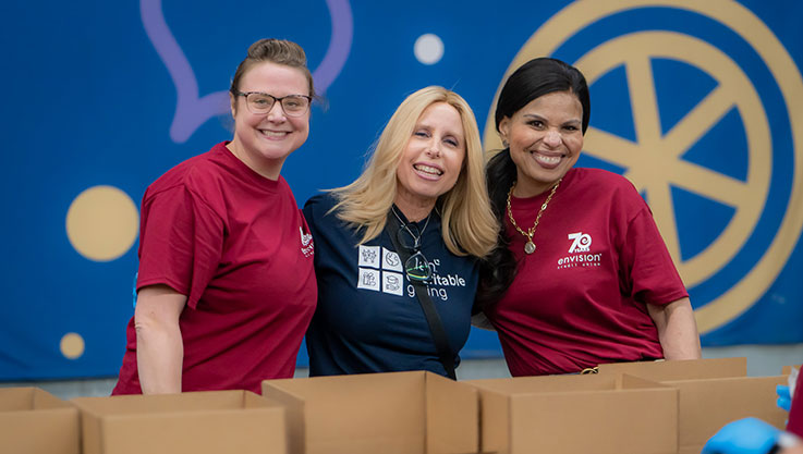 Three volunteers smile at the camera while packing boxes on an assembly line.