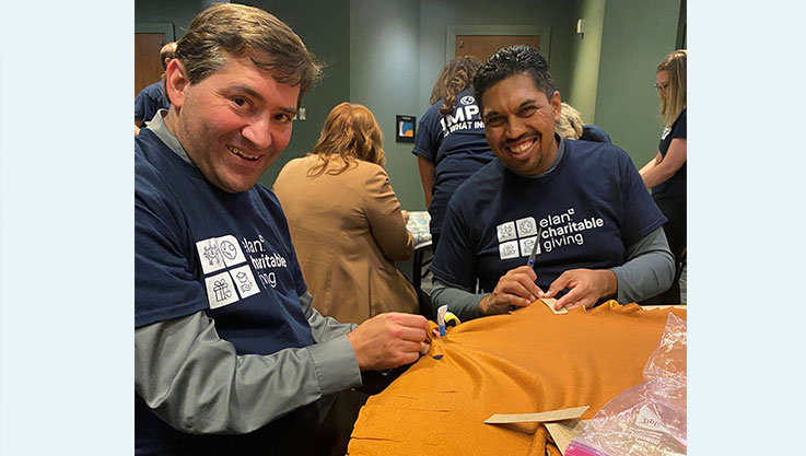 Two adults sit at a table cutting orange fabric to make a blanket. 