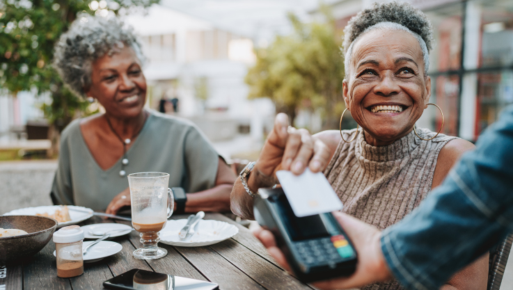 Two older women sit at an outdoor restaurant table. The woman closest to the camera holds her credit card over a tableside terminal held by the server.