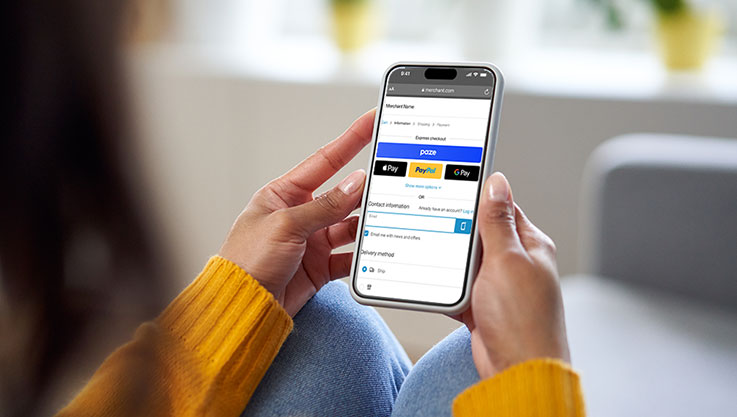 A person wearing a yellow sweater sits on a couch holding a phone. The phone screen shows Paze on the checkout page.
