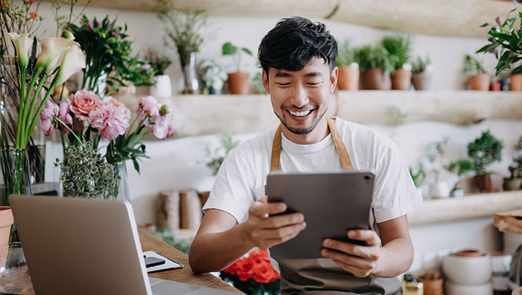 A person stands at the counter of a floral shop holding a tablet. 