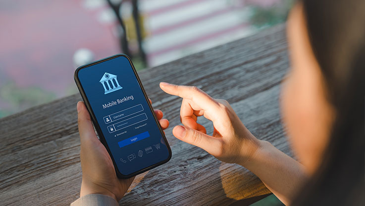 A person holds a mobile phone at an outdoor table. The phone screen shows the login screen of a mobile banking app. 