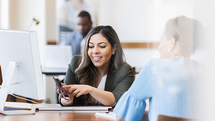 Two people sit at a desk facing each other. The woman facing the camera holds up a mobile device showing the customer the screen. 